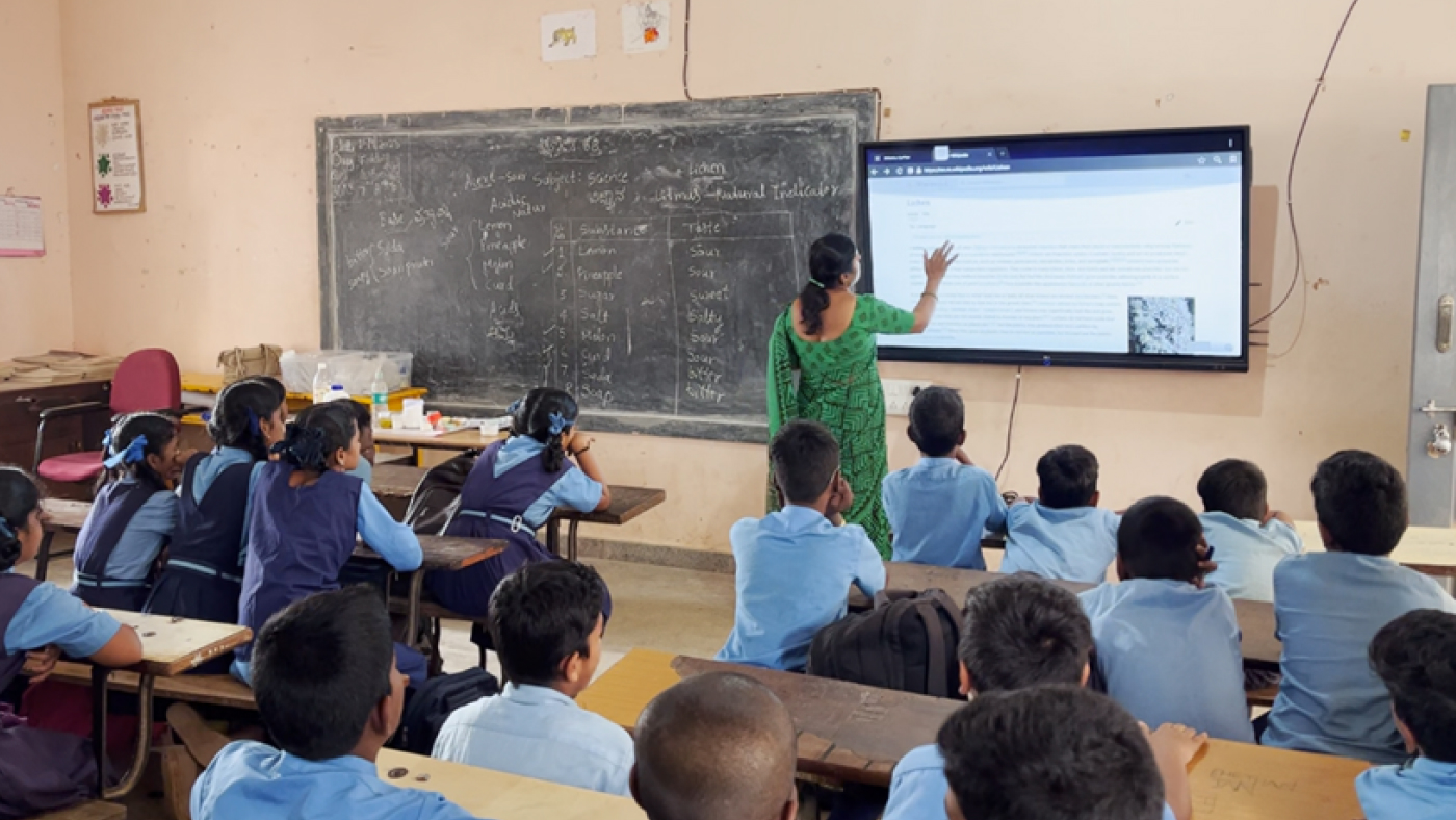 A teacher uses a classroom display while children in blue school uniforms follow the lesson